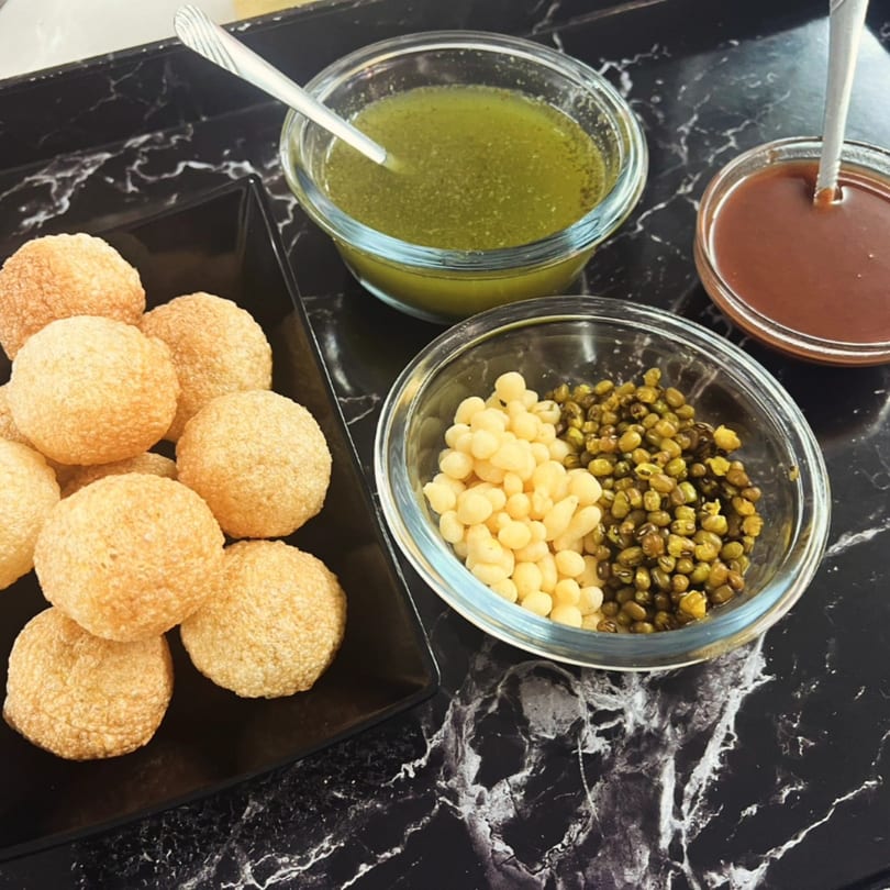 Pani Puri with green and brown sauces, and bowls of sprouts and boondi on a black marble surface.
