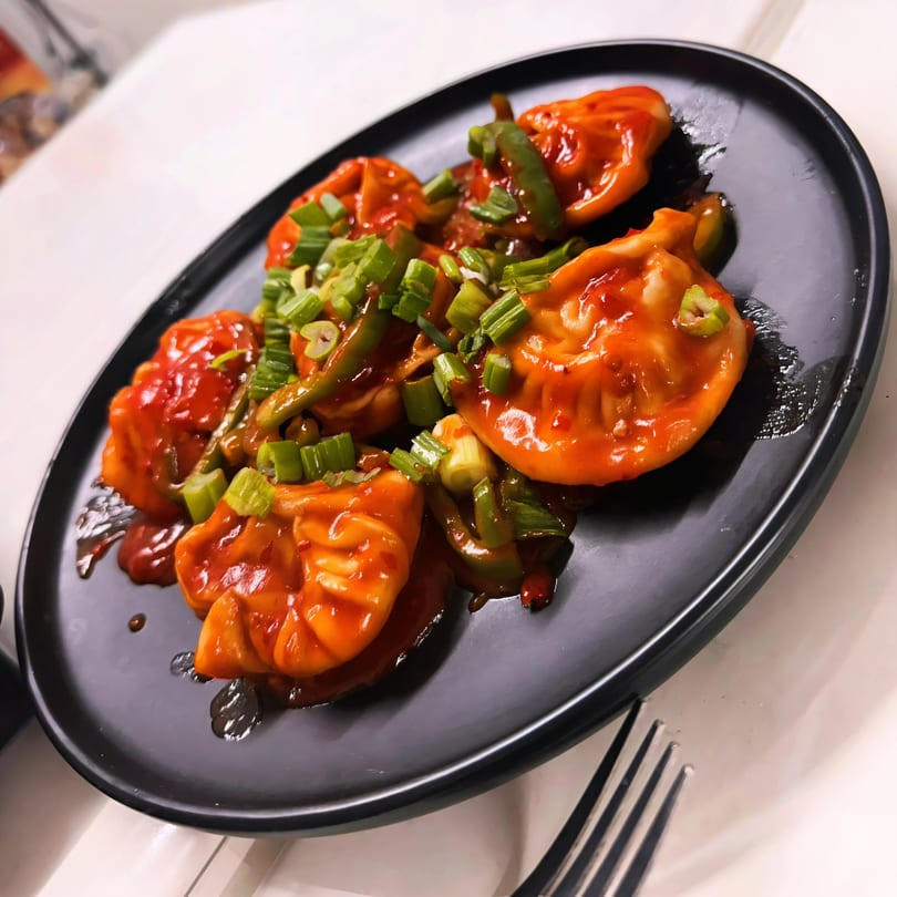 Spicy red momos topped with green onions and peppers on a black plate.